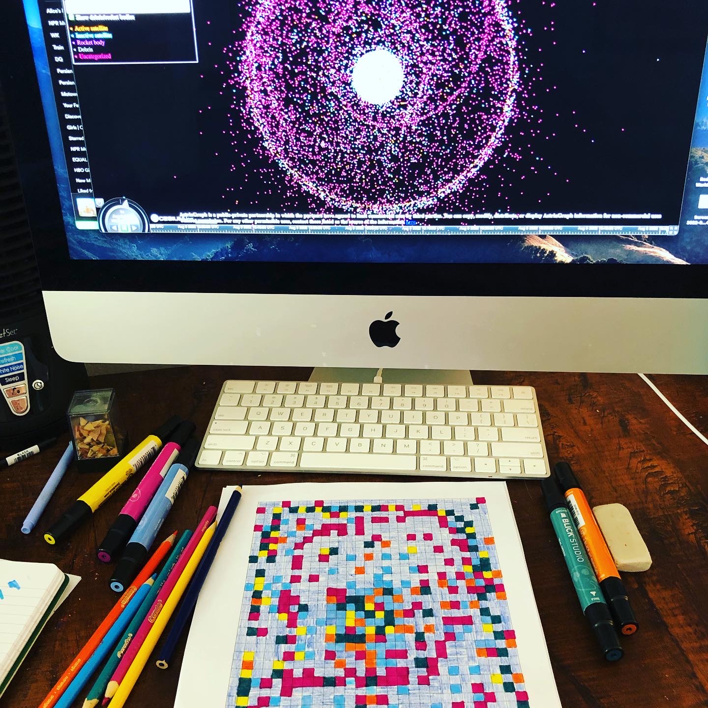 Un trozo de papel cuadriculado rellenado con diferentes colores brillantes se muestra junto a una pantalla de ordenador que muestra una representación de la basura espacial en forma de pequeños puntos en un círculo.