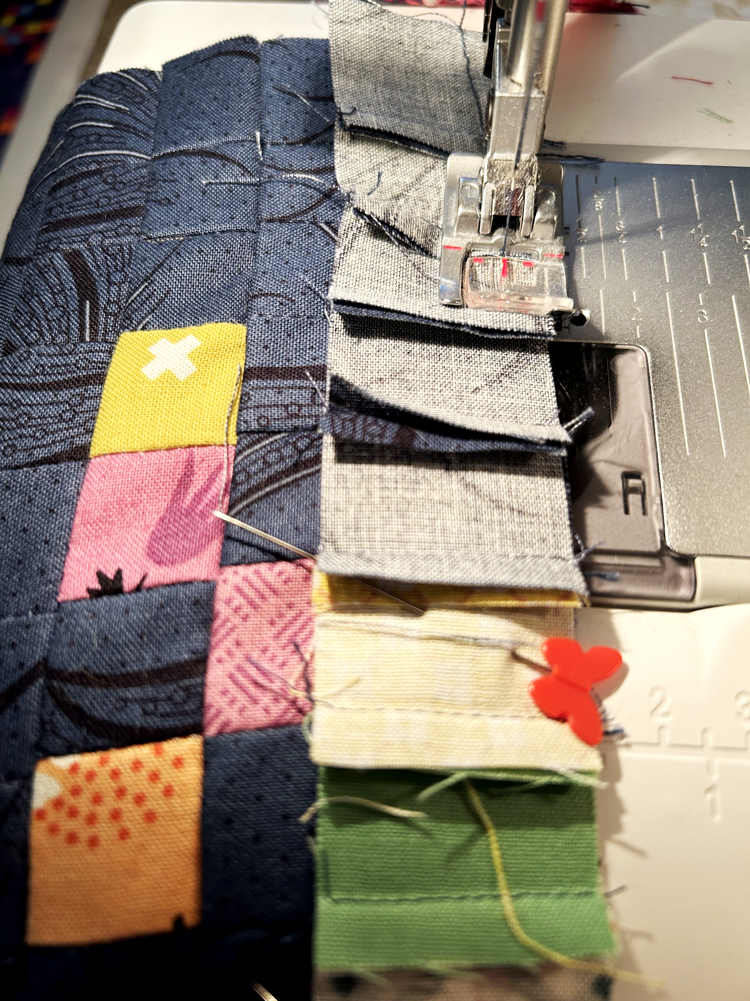 Small squares of fabric (dark blue, yellow, pink, and orange) are pinned together in a row and sewn together with a sewing machine.