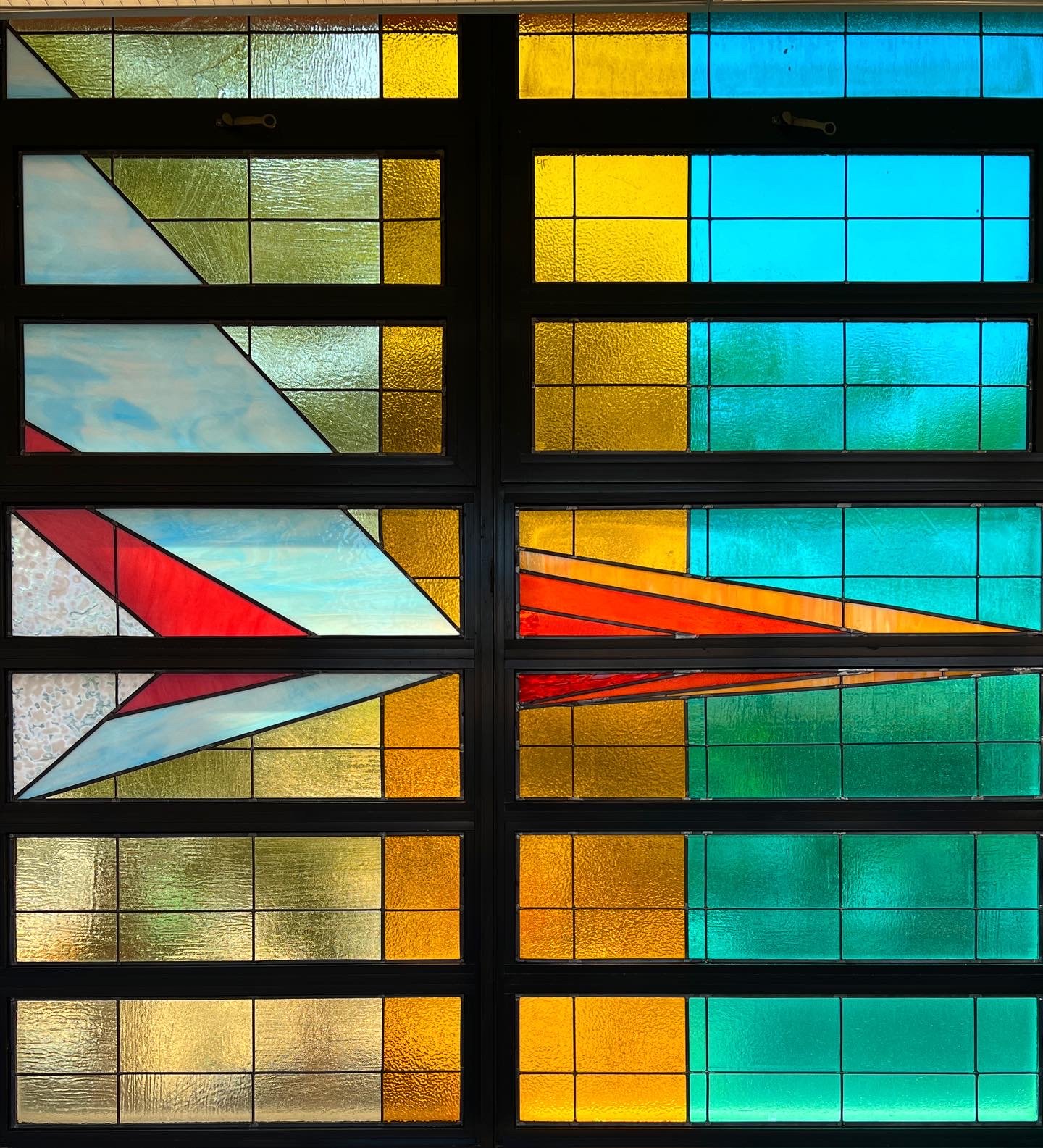 A side-view of a multi-colored, multi-paneled, stained glass window taken from the hallway of a school. Also, included are two close-ups of sections of the stained glass installation.
