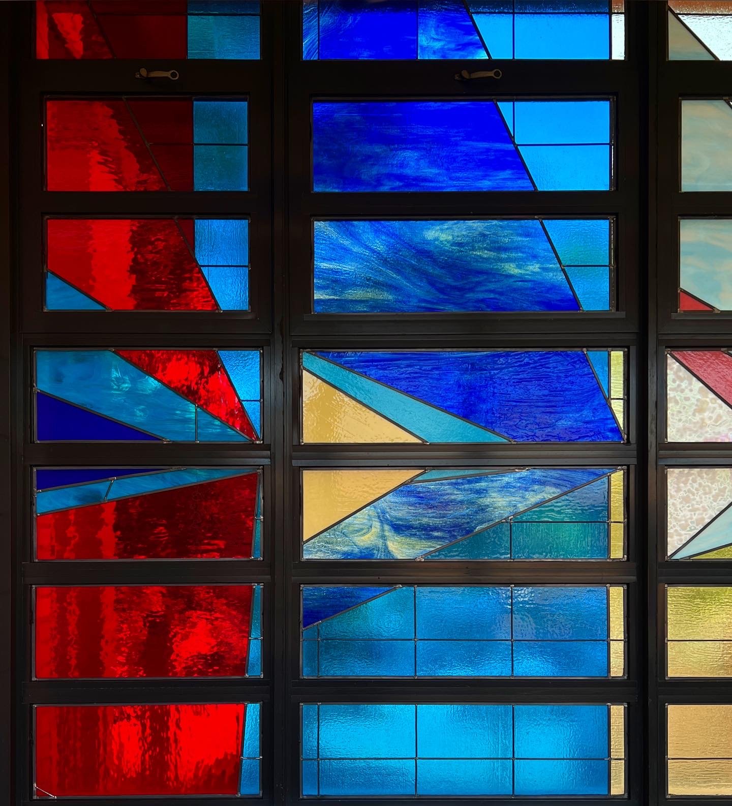 A side-view of a multi-colored, multi-paneled, stained glass window taken from the hallway of a school. Also, included are two close-ups of sections of the stained glass installation.