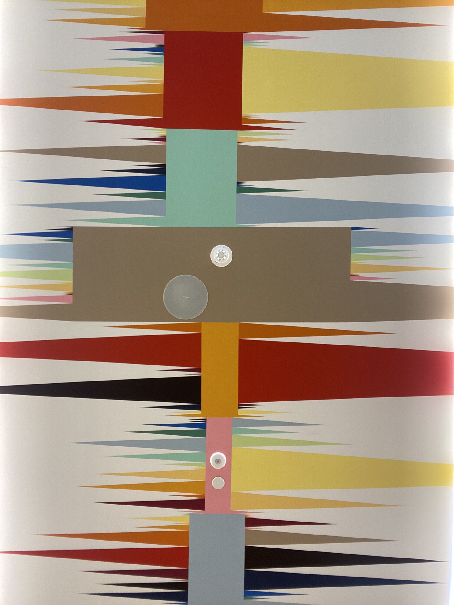 A zoomed-in view of a colorful mural painted on a white ceiling. Rectangular boxes Bars of varying widths in sevenix different colors of varying dimensions extend vertically along the central axis of the image. Both the second and fourth bars from top to bottom contain a couple of small circles representing additional information. From either side of the bars, along the horizontal axis emerge non-overlapping triangles of different sizes and colors.