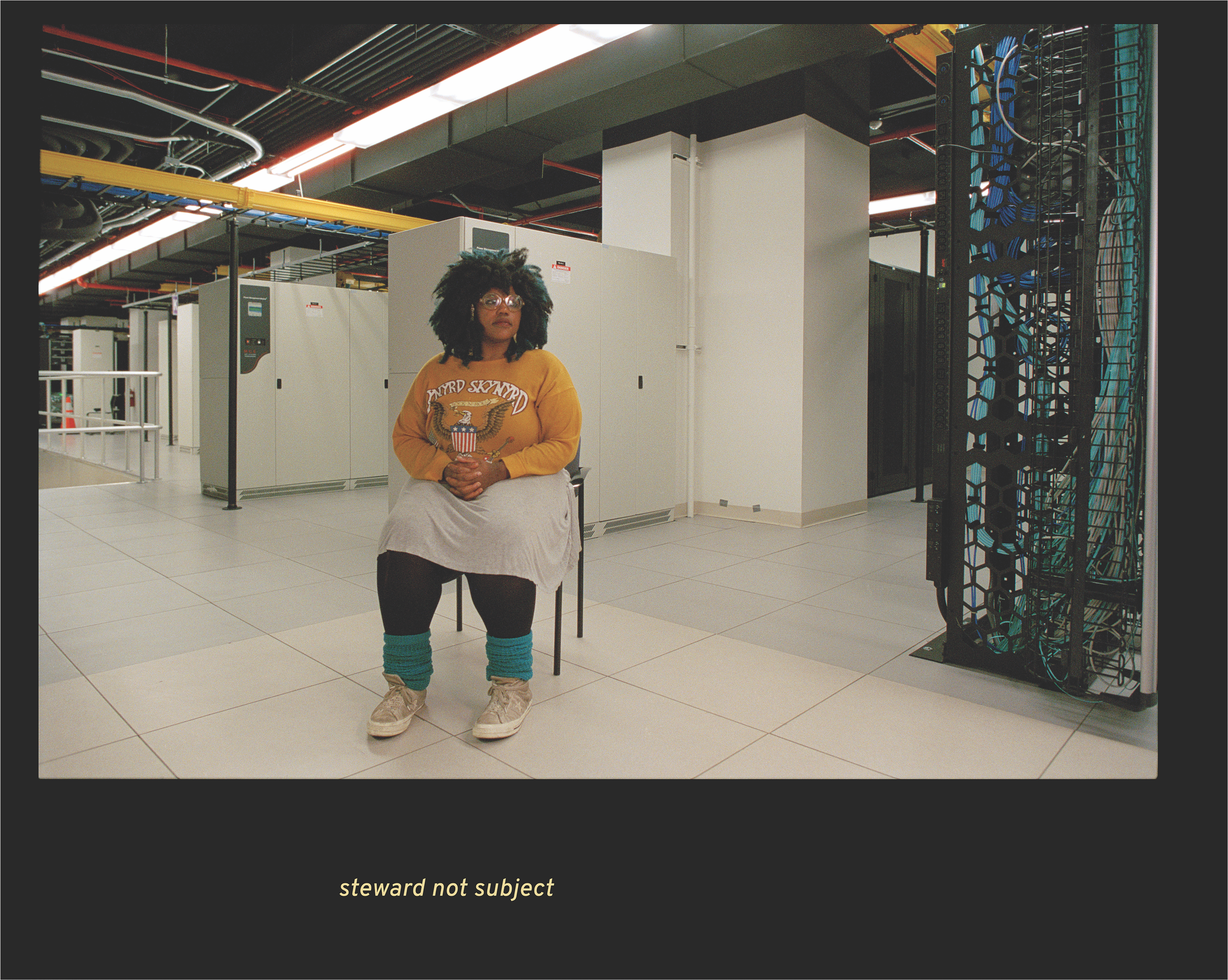 Black woman with black and blue hair sits in front of room holding electronics and metal cabinets with the caption “steward not subject”