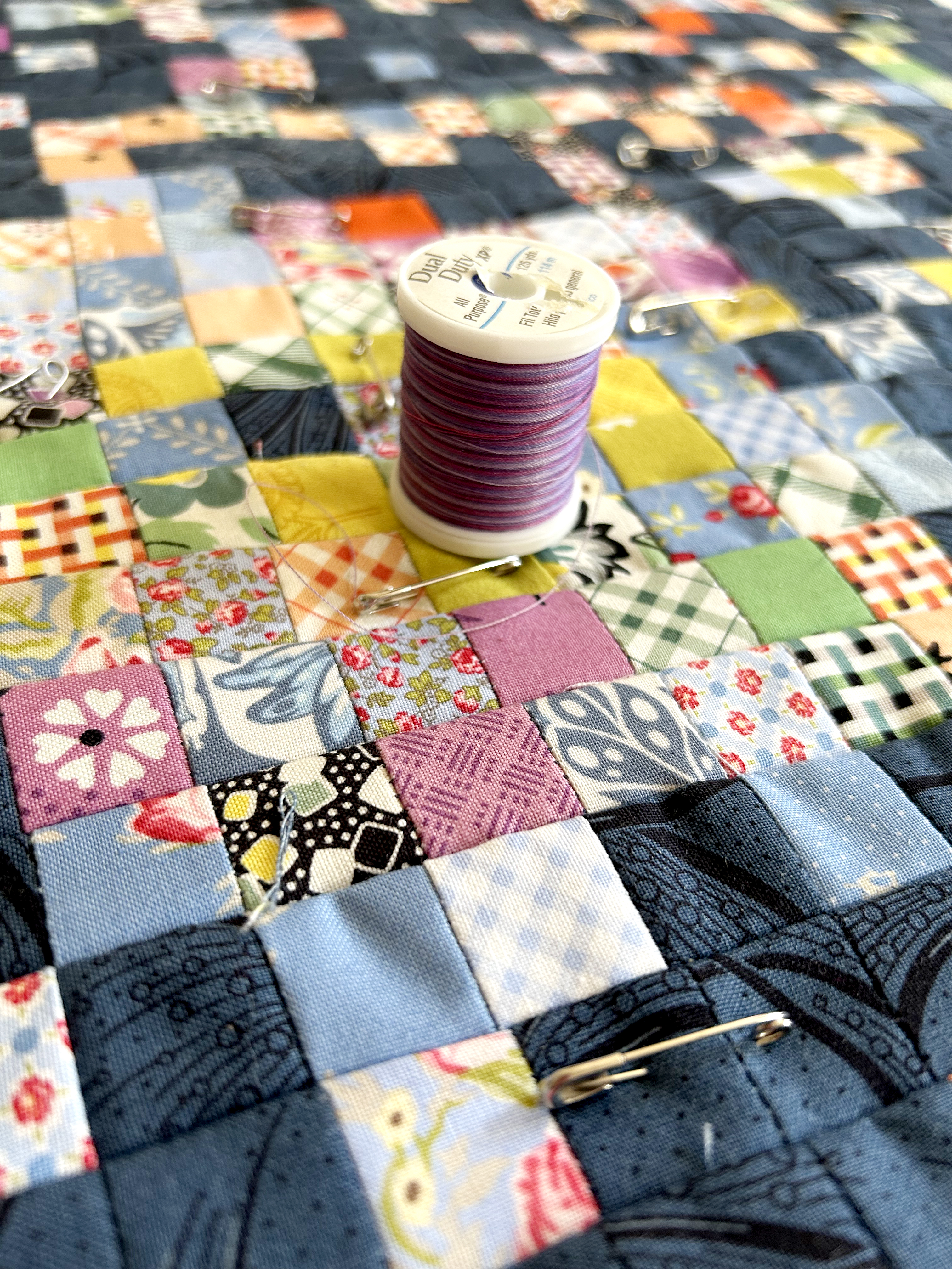 A multicolored quilt made up of small squares has been pinned with safety pins, and a pink-purple spool of thread sits on top of the quilt.