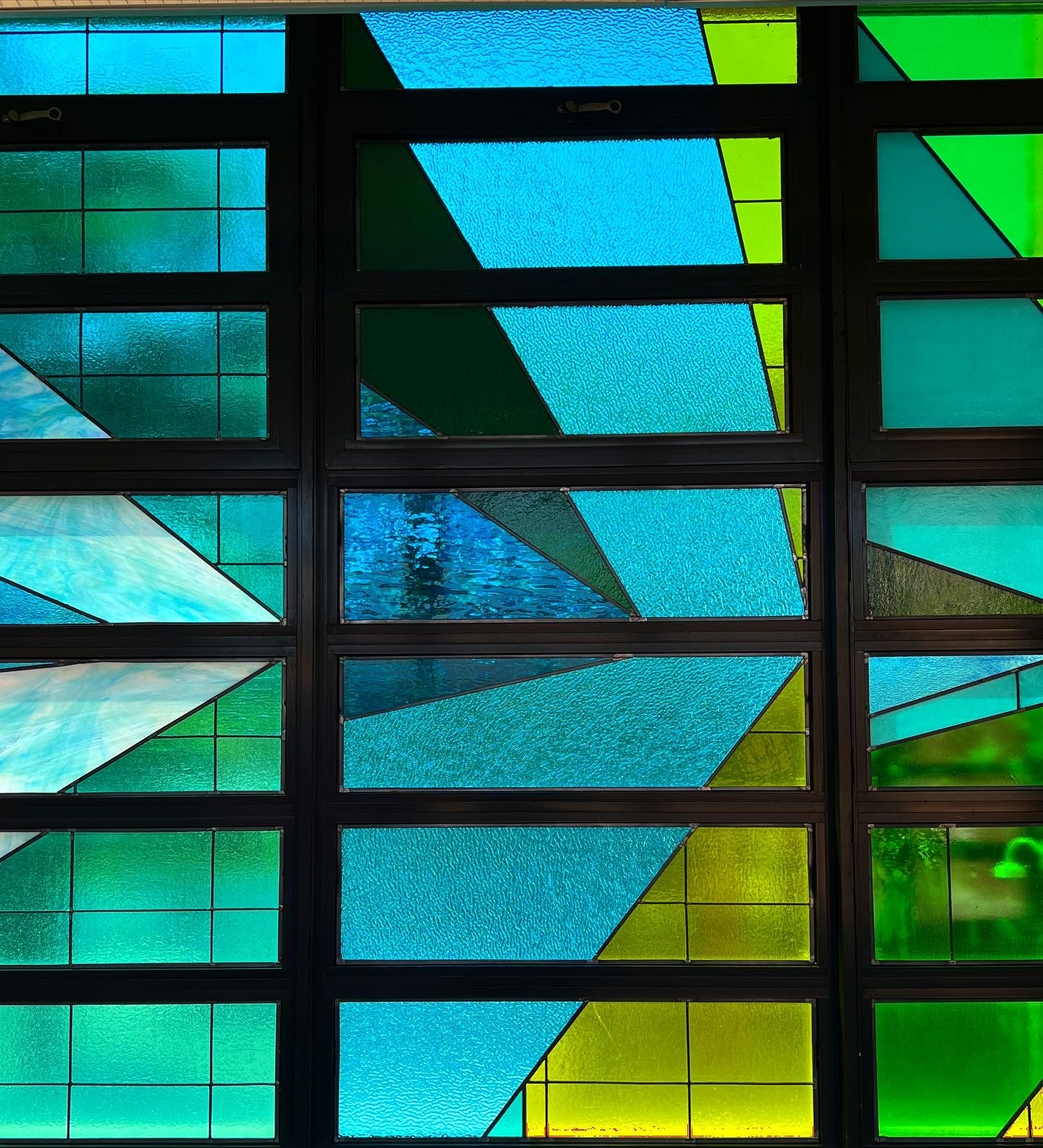 A side-view of a multi-colored, multi-paneled, stained glass window taken from the hallway of a school. Also, included are two close-ups of sections of the stained glass installation.