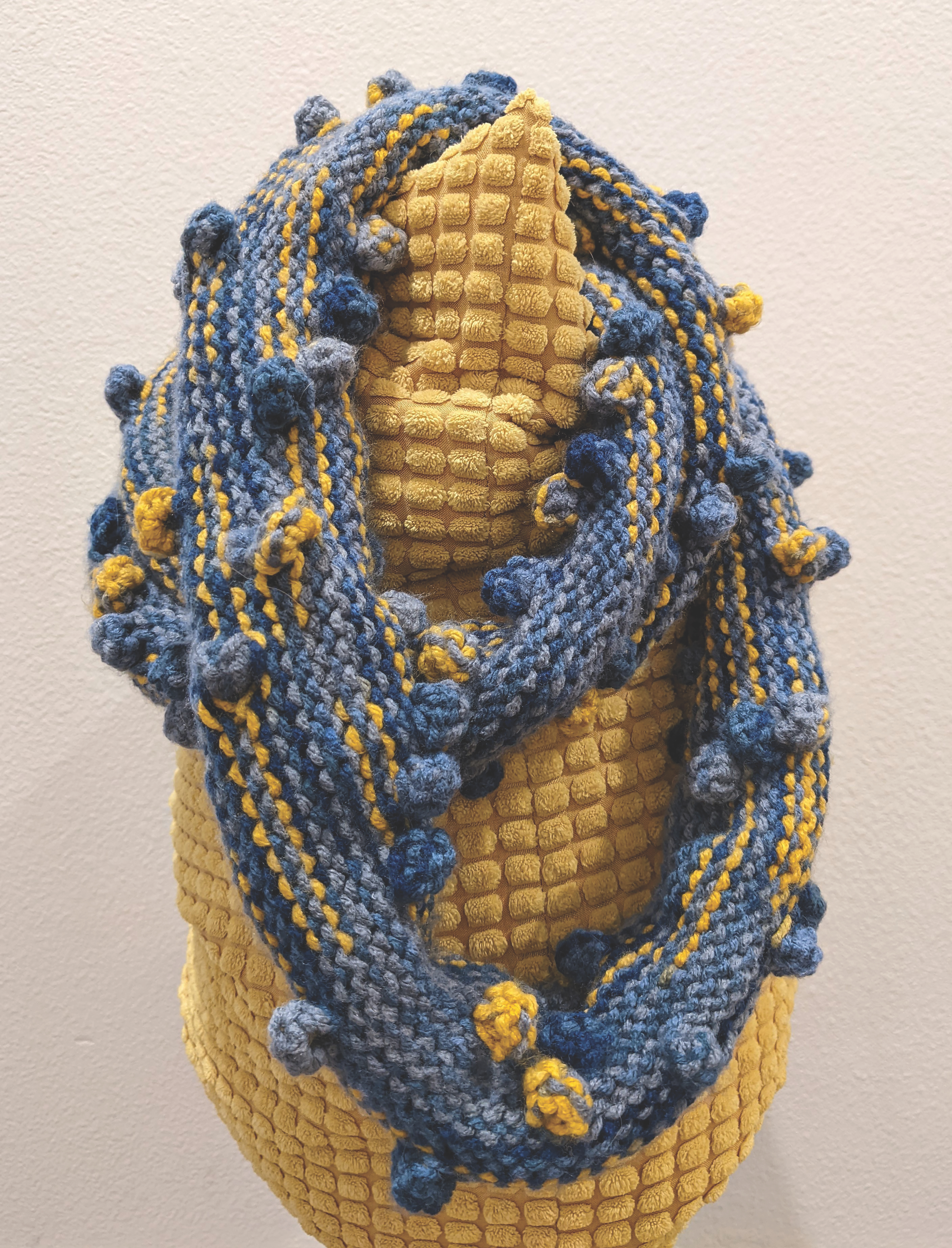 A scarf with bobbles, knitted with blue and yellow yarn, draped on a yellow cushion.